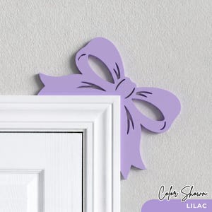 May include: A lilac-colored decorative bow cutout designed to be placed on the corner of a door frame. The bow features cut-out details and is positioned at the top right corner of a white door frame against a light gray wall.