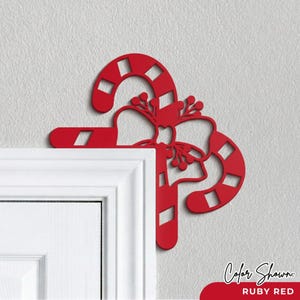 May include: A ruby red candy cane door corner decoration featuring a cutout striped pattern and a central bow with berry accents. The flat, decorative piece is positioned to fit snugly against the upper corner of a white door frame.