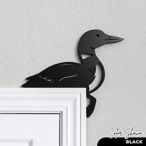 May include: A matte black metal wall art piece shaped like a loon bird, designed to be mounted on the corner of a door frame. The silhouette features cut-out details representing the bird's wing and body feathers, with a sharp, pointed beak.