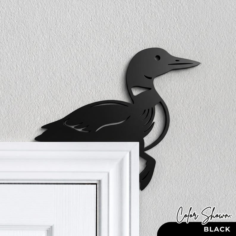 May include: A matte black metal wall art piece shaped like a loon bird, designed to be mounted on the corner of a door frame. The silhouette features cut-out details representing the bird's wing and body feathers, with a sharp, pointed beak.