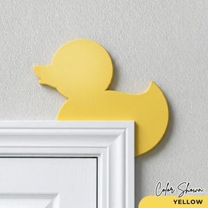 May include: A bright yellow duck-shaped doorstop. The doorstop is placed on top of a white door frame. The color shown is yellow. The doorstop is a decorative item.