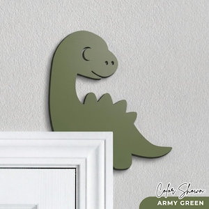 Puede incluir: Decoraci&oacute;n de pared de dinosaurio verde militar con contorno negro. El dinosaurio tiene un cuello curvado, una cara sonriente y una fila de p&uacute;as en la espalda. El texto "Color Shown: ARMY GREEN" est&aacute; en la parte inferior.