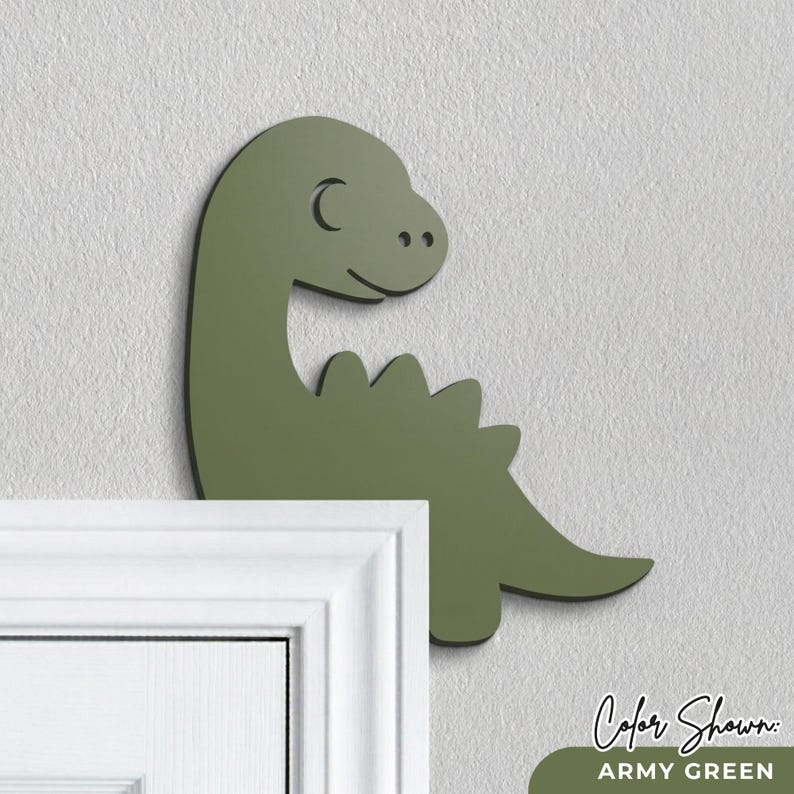 Puede incluir: Decoraci&oacute;n de pared de dinosaurio verde militar con contorno negro. El dinosaurio tiene un cuello curvado, una cara sonriente y una fila de p&uacute;as en la espalda. El texto "Color Shown: ARMY GREEN" est&aacute; en la parte inferior.