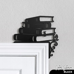 May include: A black metal door corner accent piece featuring a stack of four books. The design includes a decorative scrollwork flourish extending from the bottom right side of the book stack, intended for placement on the corner of a door frame.
