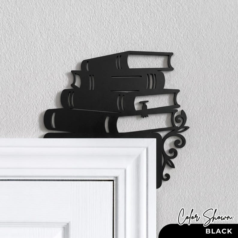 May include: A black metal door corner accent piece featuring a stack of four books. The design includes a decorative scrollwork flourish extending from the bottom right side of the book stack, intended for placement on the corner of a door frame.