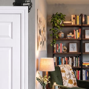 May include: A black metal door corner accent featuring a stack of books design. The decorative piece is mounted on the upper corner of a white door frame, showcasing a detailed silhouette of stacked volumes with ornate scrollwork at the base.