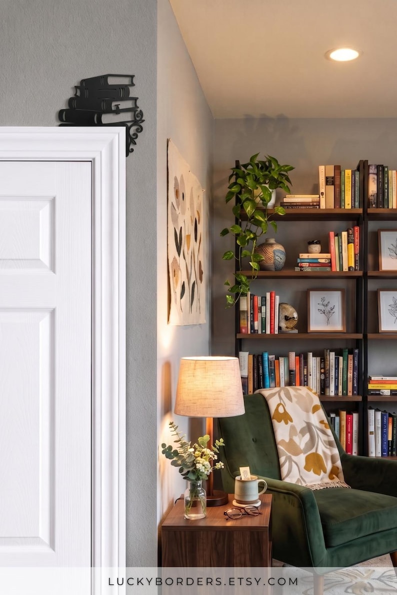 May include: A black metal door corner accent featuring a stack of books design. The decorative piece is mounted on the upper corner of a white door frame, showcasing a detailed silhouette of stacked volumes with ornate scrollwork at the base.