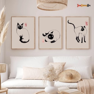Op de afbeelding: Drie zwart-witte inkttekeningen van Siamese katten in verschillende poses. De tekeningen zijn ingelijst en hangen aan een witte muur boven een witte bank.