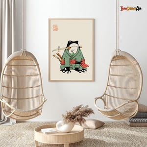 Peut inclure: Une affiche encadrée représentant une grenouille de dessin animé habillée en samouraï, tenant une épée dans sa bouche et deux autres à ses côtés. La grenouille porte un kimono vert avec des détails rouges. Le fond est beige clair.