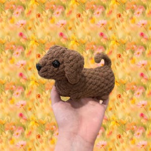 Könnte beinhalten: Ein brauner gehäkelter Dackel mit großen schwarzen Augen und einem eingerollten Schwanz. Das handgefertigte Stofftier wird in einer Hand gehalten, vor einem verschwommenen floralen Hintergrund. Der Hund ist aus weichem, strukturiertem Garn gefertigt.