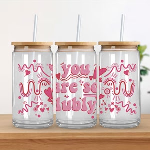 Può includere: Tre bicchieri di vetro trasparente con coperchi in bambù e cannucce rosa. Ogni bicchiere ha un design rosa e bianco con il testo "you are so lubly" sul bicchiere centrale.