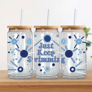 Può includere: Tre bicchieri di vetro trasparente con coperchi in bambù e cannucce. Ogni bicchiere ha un design astratto blu e bianco con cerchi e linee. Il bicchiere centrale ha il testo "Just Keep Swimming" in blu.