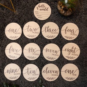 May include: A set of 12 wooden baby milestone discs with a brown wood finish. The discs feature the words "hello world" and numbers one through twelve, with the words "months" or "year" printed below each number. The discs are perfect for capturing a baby's growth and development.