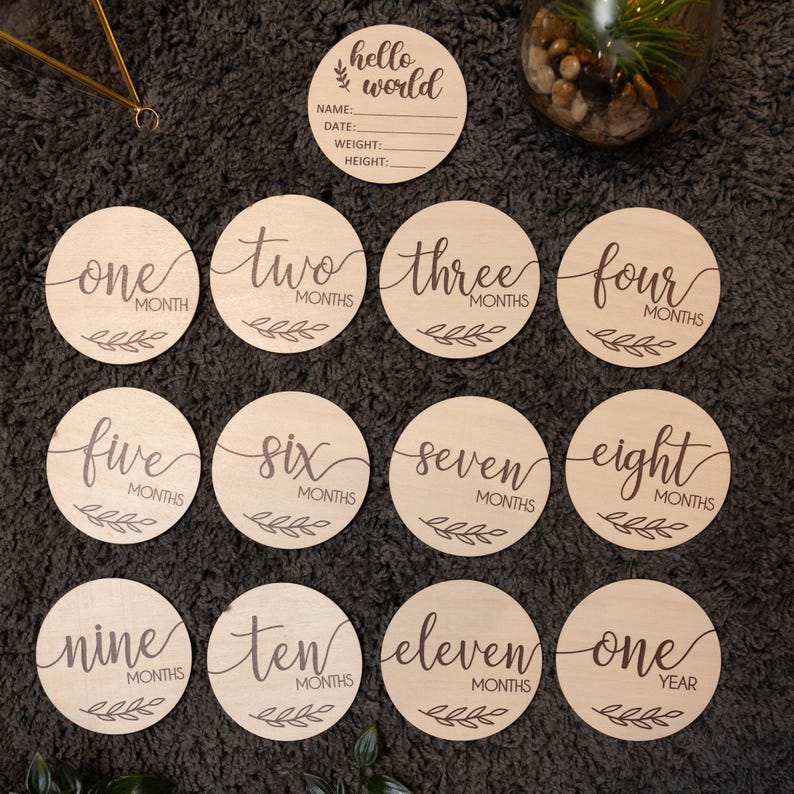 May include: A set of 12 wooden baby milestone discs with a brown wood finish. The discs feature the words "hello world" and numbers one through twelve, with the words "months" or "year" printed below each number. The discs are perfect for capturing a baby's growth and development.