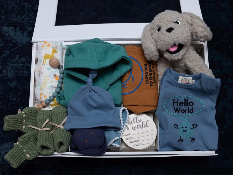 May include: A white gift box filled with baby items, including a blue and green hooded sweater, a blue hat, a blue onesie with the text "Hello World" in a speech bubble, a brown and blue blanket with the text "I love you to the moon and back", a green and brown knitted sock, and a stuffed gray dog.