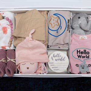 May include: A white gift box filled with baby items, including a pink and white onesie with the text "Hello World", a pink and white hat, a gray elephant stuffed animal, a pink and white blanket, a brown hooded sweater, a pink and white swaddle, and a wooden tag with the text "hello world".