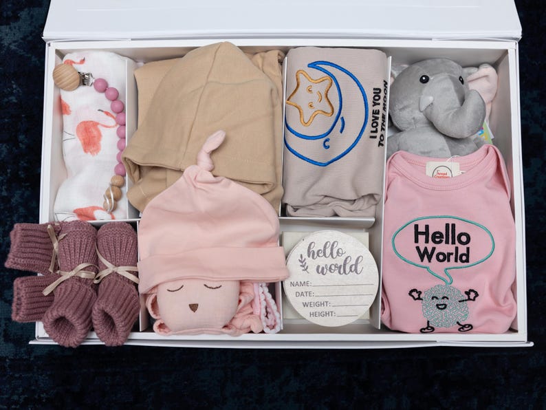 May include: A white gift box filled with baby items, including a pink and white onesie with the text "Hello World", a pink and white hat, a gray elephant stuffed animal, a pink and white blanket, a brown hooded sweater, a pink and white swaddle, and a wooden tag with the text "hello world".