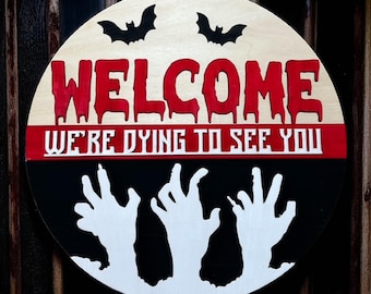 May include: Round wooden Halloween sign with the word "WELCOME" in dripping red letters. The sign also says "WE'RE DYING TO SEE YOU" and features white zombie hands reaching up from a black background, with two black bat silhouettes.
