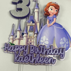 May include: Purple and silver cake topper with a princess character, a castle, and the words "Happy Birthday Kathleen" and the number "3".