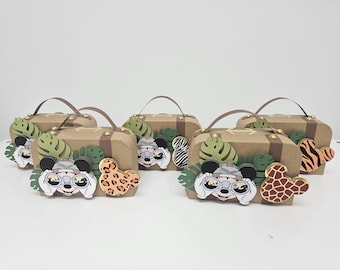 May include: Five brown cardboard boxes shaped like suitcases with gold accents. Each box has a different animal design on the front, including a zebra, a leopard, a giraffe, and two Mickey Mouse characters wearing explorer hats and binoculars. The boxes are decorated with green leaves and have gold handles.