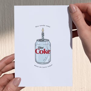 May include: A greeting card featuring a Diet Coke can with a lit birthday candle on top. The can is light blue with the red 'Diet Coke' logo. The text above reads 'Who needs cake' and below 'when we have coke?' The card is held by two hands.
