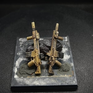 May include: Two small, detailed, black and tan toy rifles on a black display stand with a gray, textured surface.