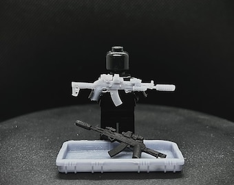 May include: A black Lego figure holding a light gray rifle with a scope and silencer. A black rifle with a scope and silencer rests in a light gray tray. The background is dark.