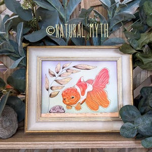 May include: A framed illustration of a goldfish with orange and white coloring. The goldfish is facing left, with a small rock and a leafy branch in the background. The frame is white with a gold trim. The text "NATURAL MYTH" is at the top.