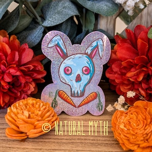 May include: A holographic sticker featuring a blue skull-shaped bunny with pink eyes and ears, and orange carrots. The sticker has a glittery effect and the text "NATURAL MYTH" is at the bottom. The sticker is surrounded by orange and red flowers.