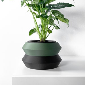 May include: A green and black ceramic planter with a leafy green plant inside. The planter has a rounded, slightly flared shape with a textured surface.