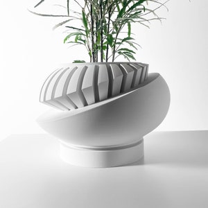 Op de afbeelding: Witte keramische plantenpot met een geometrisch ontwerp, met een groene plant met lange, dunne bladeren.