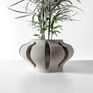 May include: A light gray ceramic planter with a unique, geometric design. The planter has a wide, round base and a narrow opening at the top. It features a series of vertical, curved lines that create a decorative pattern. The planter is filled with a green plant with long, thin leaves.