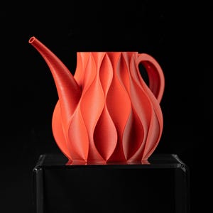 Op de afbeelding: Een levendige rode decoratieve kan met een uniek, gestructureerd ontwerp. De kan heeft een lange tuit en een handvat en wordt gepresenteerd op een transparante standaard met een zwarte basis. Het algehele ontwerp is modern en artistiek.
