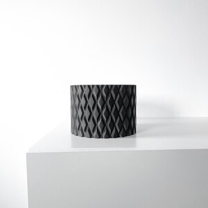 May include: A black cylindrical planter with a diamond pattern design. The planter is sitting on a white surface.
