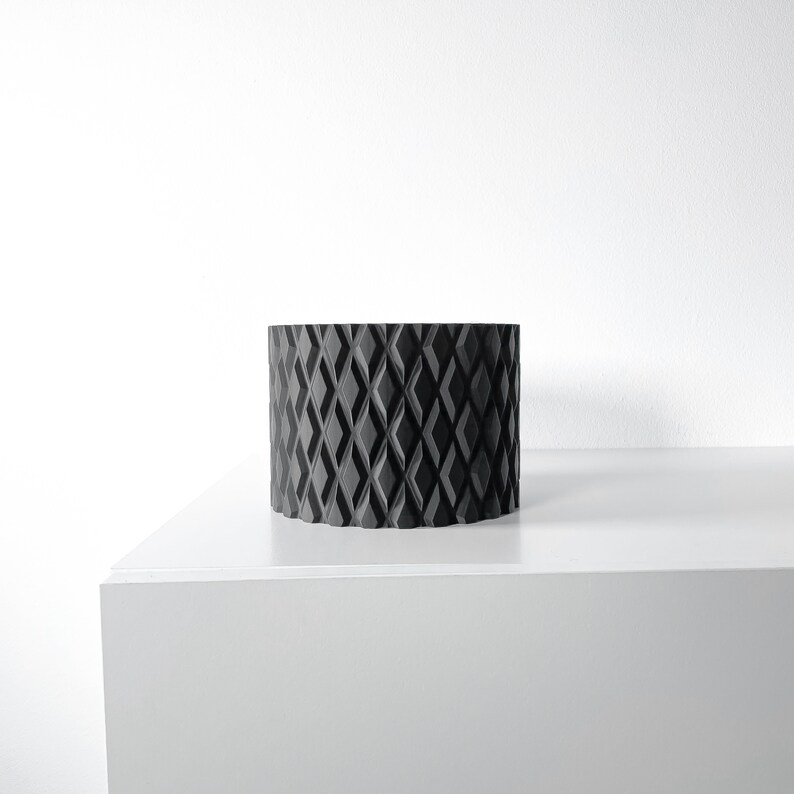 May include: A black cylindrical planter with a diamond pattern design. The planter is sitting on a white surface.