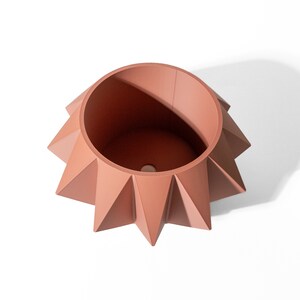 May include: A terracotta-coloured planter with a circular opening and a geometric, star-shaped exterior featuring sharp, pointed triangular ridges radiating from the centre. The vessel has a smooth matte finish and a drainage hole at the base.