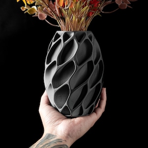 May include: A dark gray vase with an intricate, openwork design, filled with colorful artificial flowers. The vase is held in a hand against a black background, showcasing its unique texture and shape. The flowers include yellow, orange, and red.