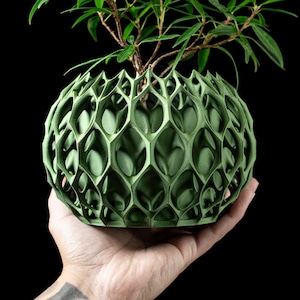 Op de afbeelding: Een groene, bolvormige, 3D-geprinte plantenbak met een geometrisch honingraatontwerp. De plantenbak bevat een kleine, groenbladige plant. De plantenbak wordt in een hand gehouden tegen een zwarte achtergrond.