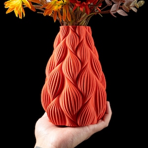 May include: A bright orange ceramic vase with a unique textured design resembling overlapping waves. The vase is being held in a person's hand.