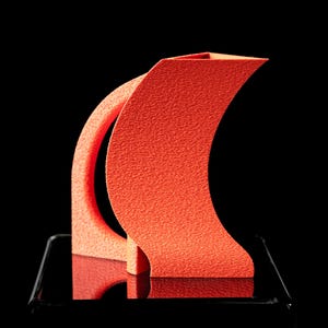Könnte beinhalten: Eine strukturierte, korallenfarbene Skulptur mit geschwungenem Design. Die Skulptur hat eine rechteckige Basis und eine kreisförmige Öffnung. Der Hintergrund ist schwarz und hebt die Form und Farbe der Skulptur hervor. Es ist wahrscheinlich ein dekoratives Kunstwerk.