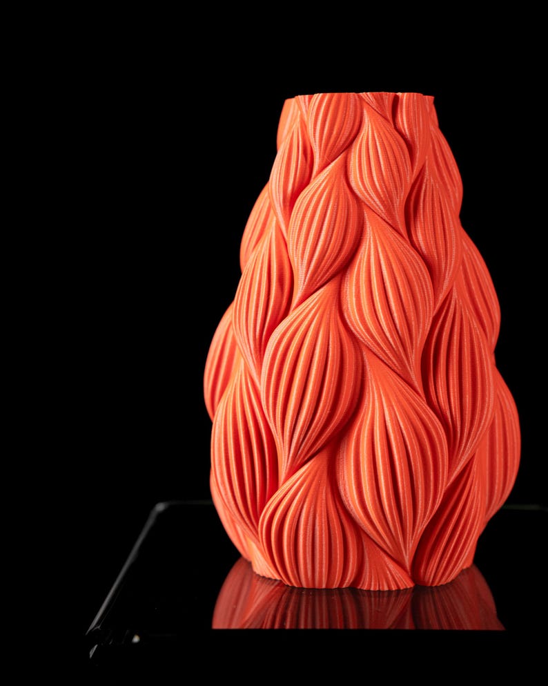 May include: A bright orange ceramic vase with a unique, textured, wavy design. The vase is shaped like a teardrop and has a smooth, glossy finish.