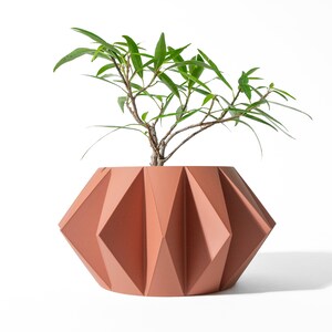 May include: A terracotta-coloured planter with a geometric, faceted, origami-inspired exterior surface. The pot features a wide, rounded base that tapers towards the top rim. A small green plant with thin, pointed leaves grows from the centre.