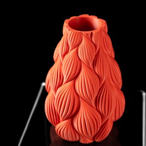 May include: A bright orange ceramic vase with a unique textured design resembling overlapping petals. The vase is on a black reflective surface.