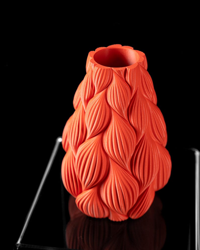 May include: A bright orange ceramic vase with a unique textured design resembling overlapping petals. The vase is on a black reflective surface.