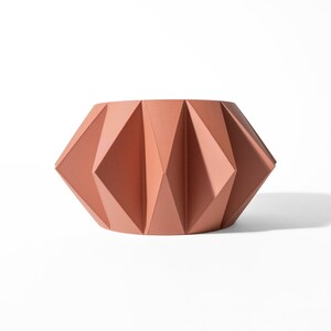 May include: A terracotta-coloured ceramic planter featuring a geometric, pleated design with sharp, angular folds that create a repeating diamond pattern around the exterior. The vessel has a wide, open top and a tapered base with a matte finish.
