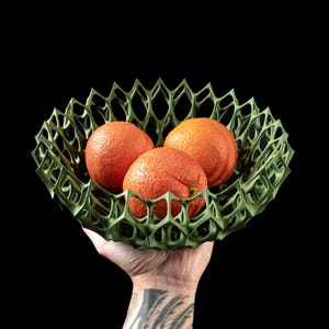 May include: A green, geometric, 3D printed bowl holds three bright orange oranges. The bowl is held in a hand with a tattoo.
