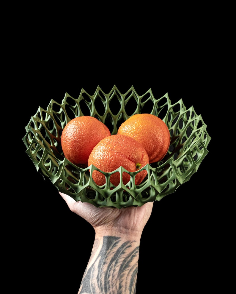May include: A green, geometric, 3D printed bowl holds three bright orange oranges. The bowl is held in a hand with a tattoo.