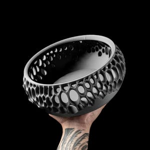 Può includere: Una ciotola in bianco e nero con un design a nido d'ape unico. La ciotola è tenuta in mano su uno sfondo nero. Il bordo della ciotola è bianco e il corpo è grigio scuro con un motivo aperto e strutturato.