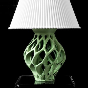 May include: A modern table lamp with a white pleated lampshade and a light green, openwork base. The base has a unique, organic design. The lamp is set against a black background, highlighting its form and texture.