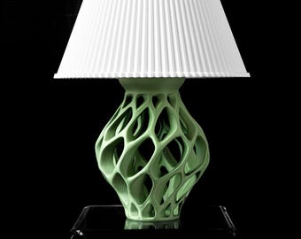 May include: A modern table lamp with a white pleated lampshade and a light green, openwork base. The base has a unique, organic design. The lamp is set against a black background, highlighting its form and texture.